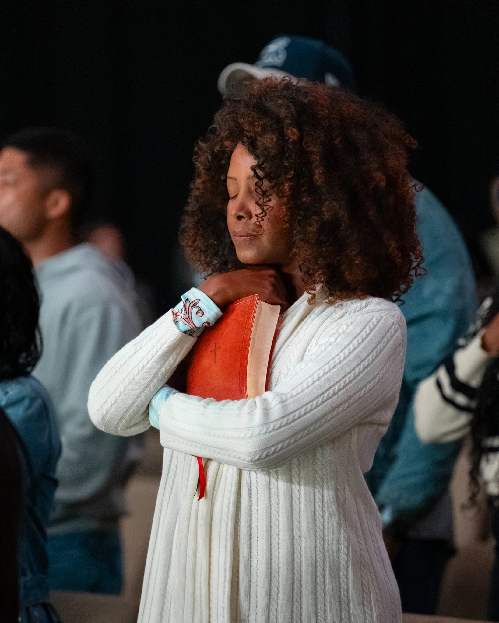 Woman in a white knit sweater holding an orange-red Bible close with her eyes closed while praying.